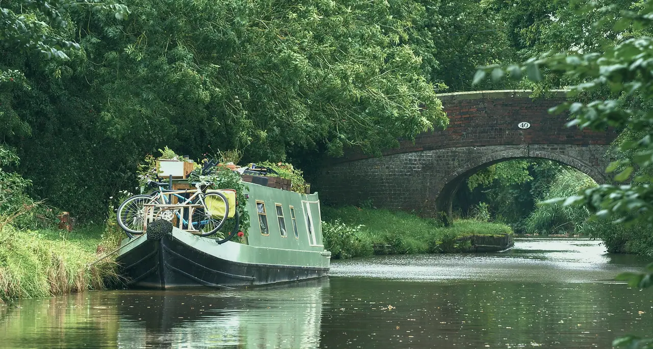 A green narrowboat with a bicycle and pots of herbs on its roof, moored below a brick bridge