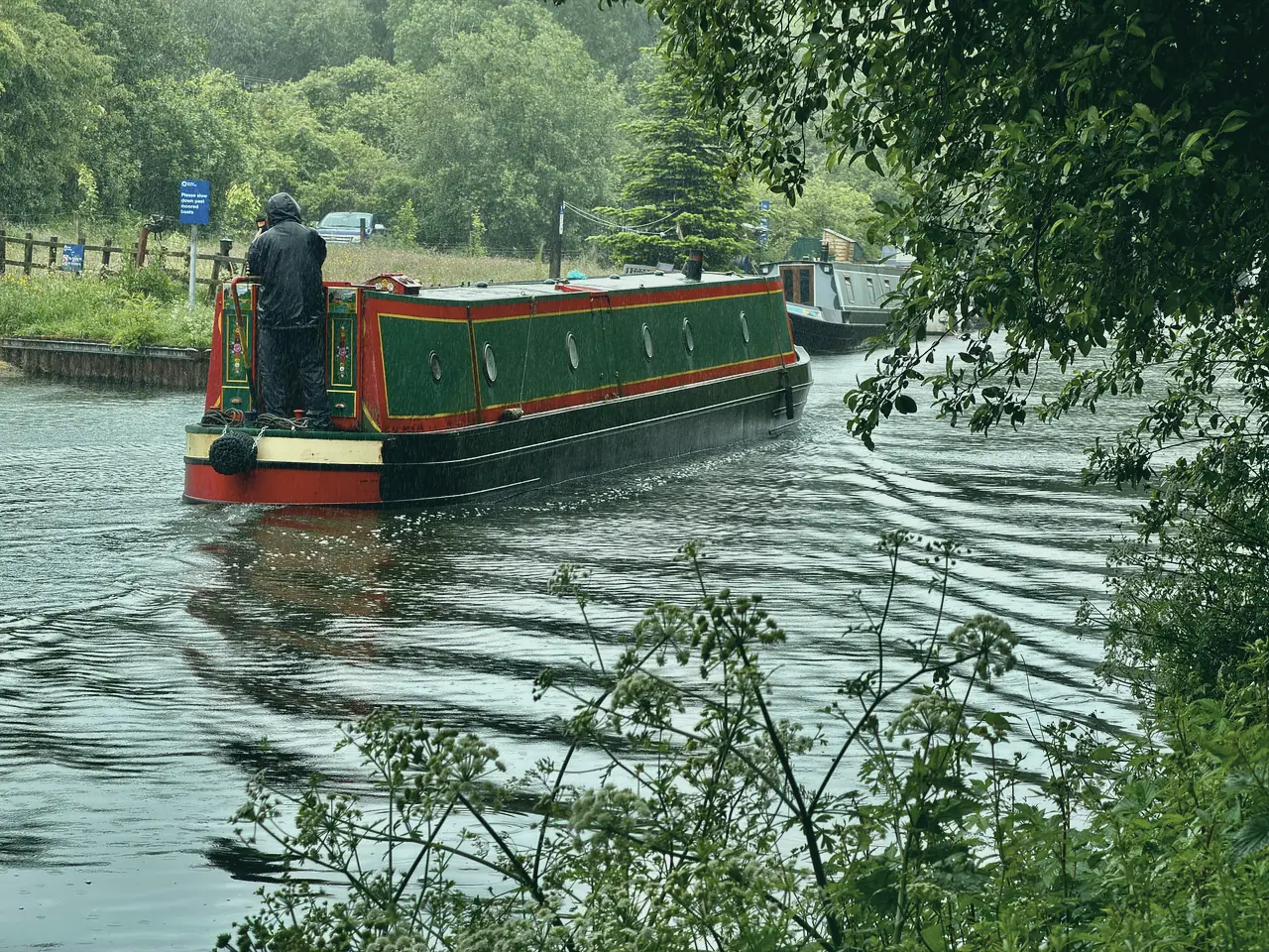 A red-and-green narrowboat cruising away down a rippling canal, the steerer at the back