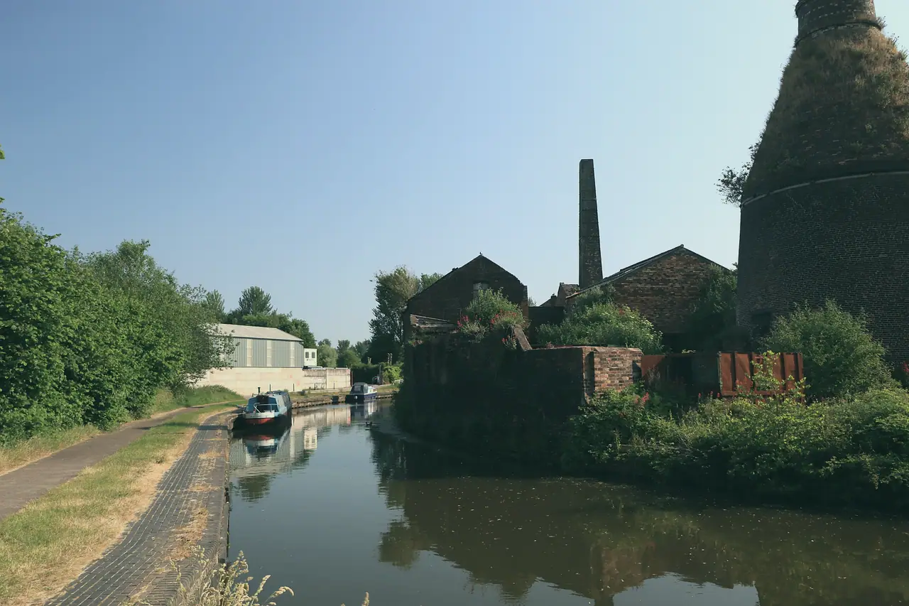 A calm canal reflecting old brick wharf buildings and a chimney under a blue sky