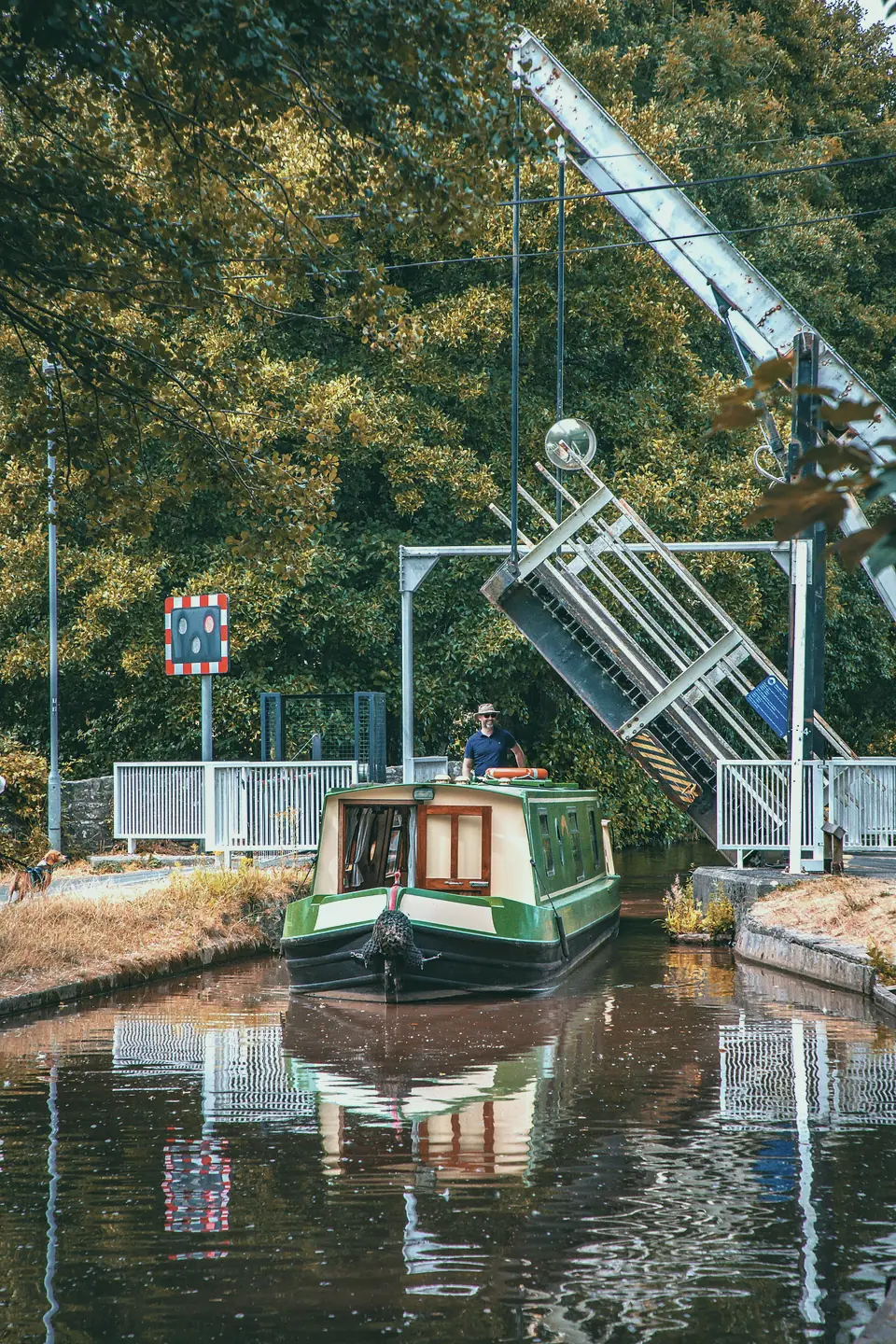 A green-and-cream narrowboat passing through a raised lift bridge, the steerer smiling at the tiller