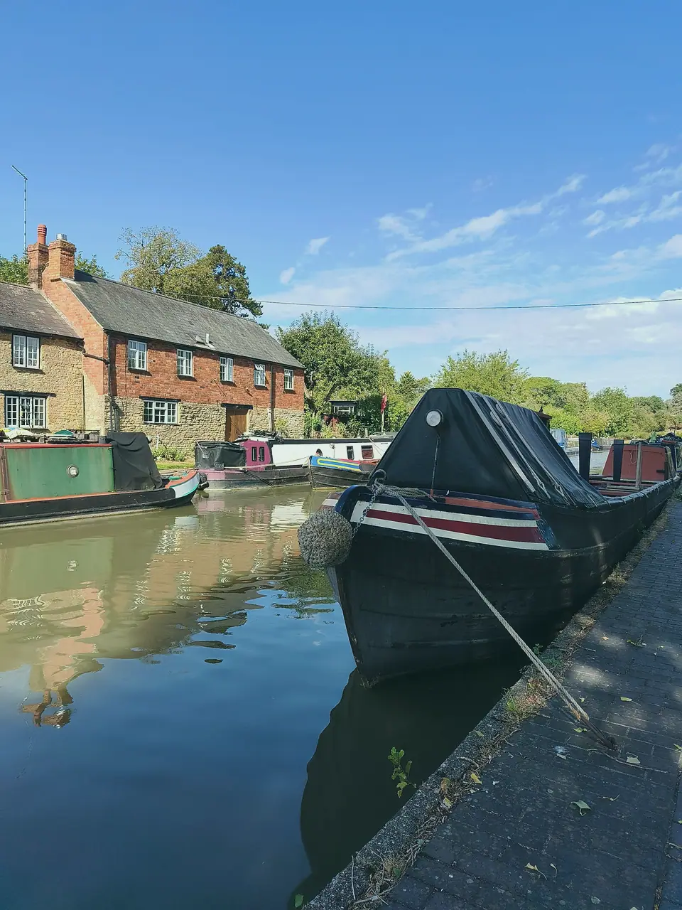 A black narrowboat with a red-and-white striped bow, moored by a brick wharf cottage